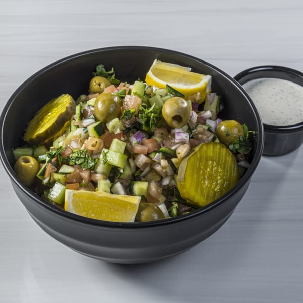 A black bowl of chopped salad with pickles, olives, and lemon wedges, with a side of dressing.